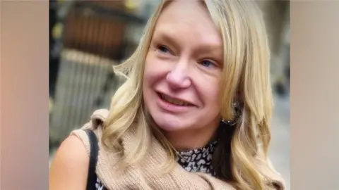 A woman with blonde hair wearing a black and white top wit a beige scarf. She is smiling and looking to the side