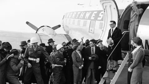 Getty Images Actors grouped around the door of a white North West airplane, which has its steps down and door open. A man in a black overcoat, white shirt and striped tie holds his bowler hat as he stands on the middle stair and looks at the crowd. Others are dressed in military uniforms with hard hats, with civilians in suits, bowler hats and overcoats. Some onlookers hold up notebooks and pens or cameras.