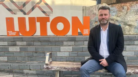 Qays Najm/BBC James Taylor wearing a dark blazer, light shirt and grey trousers sits on a wooden bench in a paved public square. Behind the bench is a large orange sign spelling “LUTON” in block letters, set against a wall featuring geometric artwork and an aged brick mural. Stone steps lead up to the sign, and surrounding buildings are visible in the background. The image is taken outdoors in daylight.