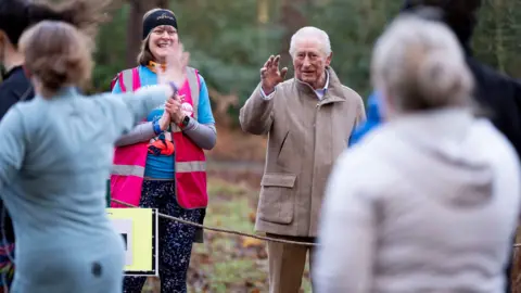 PA Media A woman standing next to King Charles, who is wearing a brown zip-up jacket and waving at a runner. There are a few runners waving at him as they are running. 