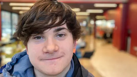 A man with brown hair smiles in the camera. He is wearing a blue hood top.