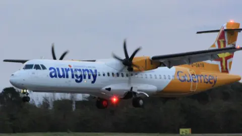 An Aurigny turboprop aircraft in yellow and white livery flies low with its landing gear down, preparing to land. The word “Aurigny” is visible on the fuselage and the tail shows a red‑and‑yellow cross design that's based on the flag of Guernsey. 