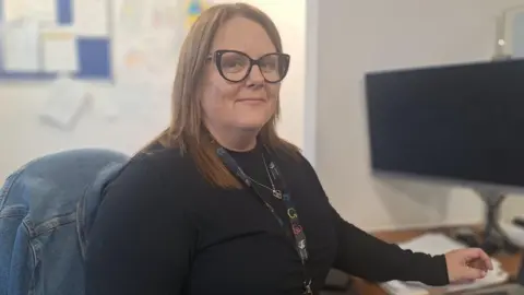 Housing officer Hannah Berry sits at her desk at the housing options drop in in Newport Central Library. She is sat on an office chair, turning to face the camera. She has a computer in front of her and a lanyard around her neck. She has long, light brown hair and large dark rimmed glasses.