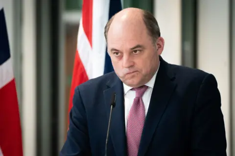 PA Media A balding man looks sternly out over a microphone, with the union jack displayed behind him. He is wearing a navy blue suit jacket, white shirt and a pink tie. 