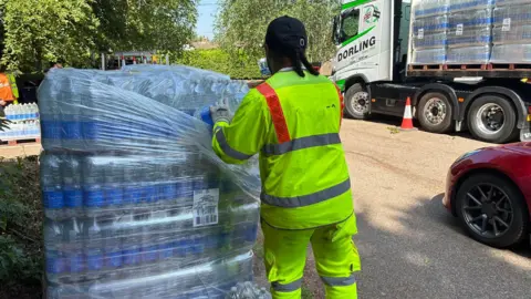 A contractor in a hi-vis jacket and trousers. He is getting slabs of water out of their shrink packaging. 