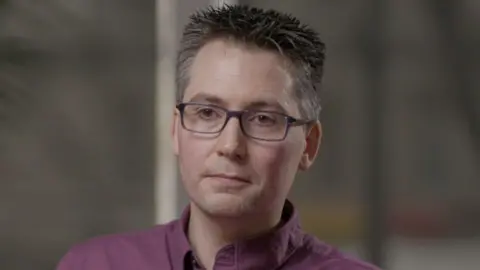 Chris Wynne has dark hair, which is grey at the sides, is wearing a purple shirt and purple, rectangle framed glasses. He is looking to the side of the camera. The background of the image is blurred. 