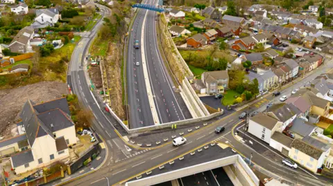 FCC Construction An aerial view of the Heads of the Valleys road and John Watkeys' house. A bridge with a road on it can be seen crossing above the dual carriageway below, with cones going down the middle of each carriageway. On the upper level, a row of terraced houses can be seen right right by the side of the road with a housing estate to the right hand side.