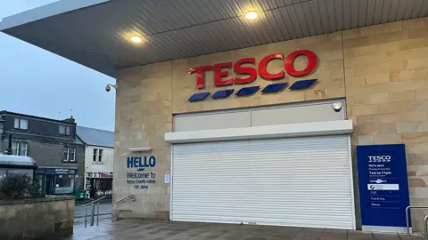 A closed Tesco in Dunfermline. The shutters, which are white, are down. There is a sign reading 'Tesco' in red lettering above.