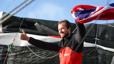 A bearded man in a sailing suit holds a union flag and in the other hand has a thumbs up.