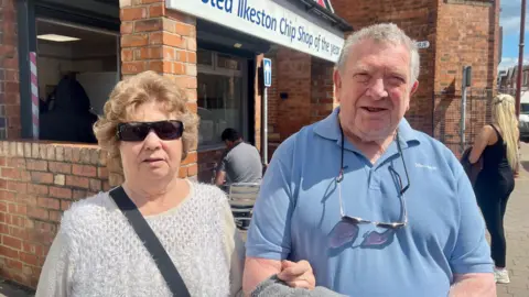 A man in a blue polo and a woman in a white top and sunglasses stand outside a fish and chip shop. There is a sign above the shop that reads "Voted Ilkeston Chip Shop of the Year."