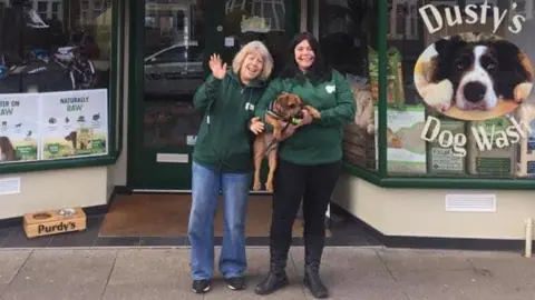 BBC Two women wearing green hooded jumpers stand outside the front entrance of a shop. The woman on the right is holding a dog and a picture of a dog is visible on the shop window.