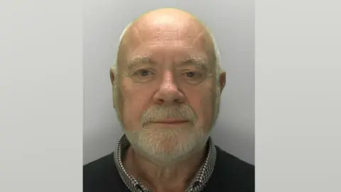 Custody image of Peter Bond. He has short white hair around the side of his head, but is bald on top. He has a white beard, and is wearing a white shirt with small blue and red checks, with a jumper over the top
