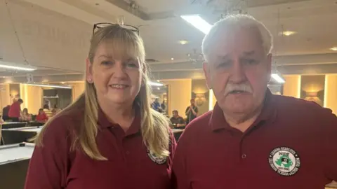 A blond haired woman and a white haired man stand beside one another. She is smiling and has glasses on her head. He has a white moustache. Both are wearing red polo shirts.