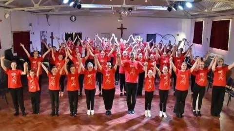 Southend Dance Centre A church hall full of dancers of different ages in red T-shirts with a logo and black trousers.