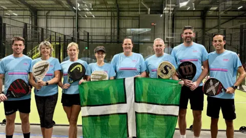 Eight people in a line wearing matching t-shirts and black shorts. They are each holding a Padel racket. the people in the middle are holding a Devon flag. They are all stood in a Padel court.