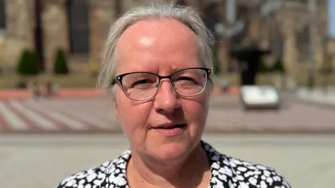 BBC/ Josh Sandiford Sarah Gayton has grey hair swept back over her head. She is wearing a pair of black-rimmed glasses and has a black blouse with a white flower pattern on it.
