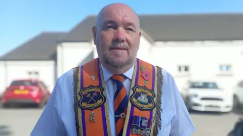 A man is wearing an orange sash with badges and medals. He has a stripped orange and purple tie on with a blue shirt. Cars are in the background and a building. 