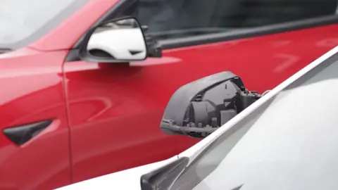 Red and white Tesla cars parked in a car park with wing mirrors damaged 