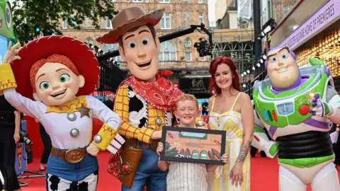 BBFC Tommy is smiling while standing on the red carpet and holding his framed design. He is standing next to a woman with red hair and they are surrounded by three people in costume as Jessie, Woody and Buzz Lightyear. 