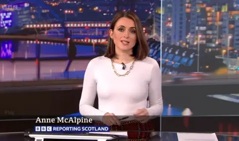 Anne McAlpine, a young woman with a rich brown bob reads the news in the Reporting Scotland studio, a large screen with a cityscape of Glasgow behind her. She wears a white top, a chunky gold necklace and a red tartan skirt. She is holding a pen.