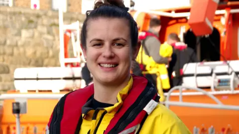 The picture shows a woman wearing a bright yellow RNLI waterproof suit and a red lifejacket with RNLI labels clearly visible. In the background, there is an orange lifeboat with crew members in similar yellow gear.
