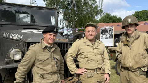 Stuart Howells/BBC Three men wearing World War Two military-style clothing smile at the camera. They stand in front of an old defender-style vehicle.