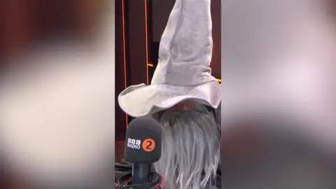 Scott Mills sits in front of a microphone. He wears a large grey wizard's hat and a long grey fake beard