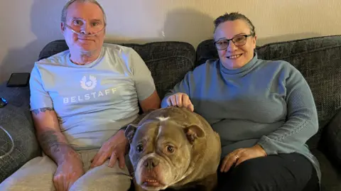 A man with a breathing tube in his nose sits next to a woman and a brown dog on a grey sofa. The couple smile at the camera. 