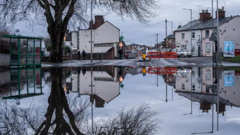 Ian Knight / Z70 Photography A large area of flooded road with a bus stop to the left. A tree is reflected in the water as well as some buildings and an orange road block. 