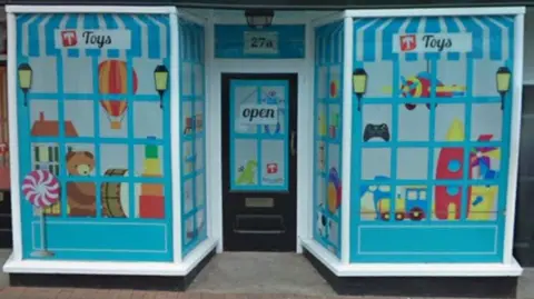 The front of one of the shops with a black door between two bay windows. The windows, with white frames, have the glass filled with a painted scene of a toy shop with musical instruments and stuff toys visible