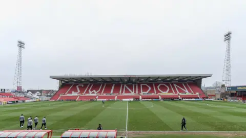 A football stadium stand with the words "Swindon" written in white letters against a red background. Stadium lights and and other buildings can be seen in the background.