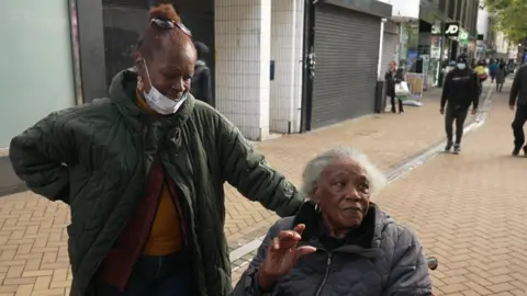 An image of a woman with dark red hair in a bun, wearing a face mask, and an older woman with short grey hair using a wheelchair, on a high street