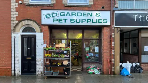 The shop front of BD Garden & Pet Supplies taken the following day after the flood water had passed. The pavement is very damp and there are sandbags visible blocking the entrance to an alley next to the shop