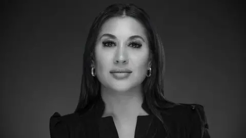 MA Legal Ltd Miriam Altaf with long dark hair and circular earrings wearing a black top looking at the camera against a black background.