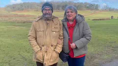 Man in black hat with glasses and beige coat stands arm in arm with woman. She is wearing a grey coat, red jumper and has grey hair.