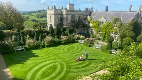 An external image of Euridge Manor, showing a freshly-mown lawn with patterns in the grass and NHS in the middle. There is a large manor house surrounded by well-pruned shrubbery.