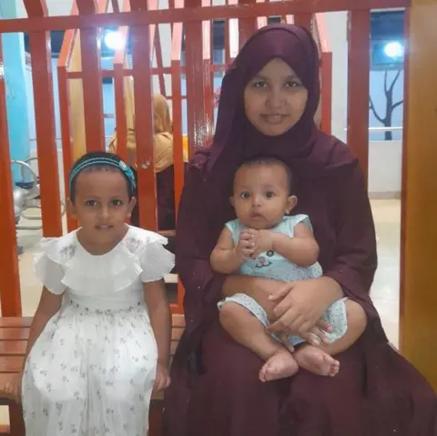 Supplied A woman wearing a maroon outfit sits on a wooden bench indoors, holding a baby in a light blue outfit, while a young child in a white dress sits beside them. Behind the bench are vertical orange bars and colourful wall decorations.