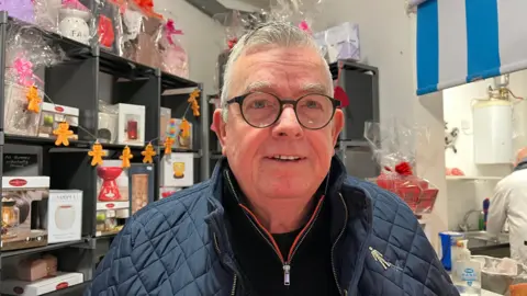 Gerry photographed at the stall, behind him you can see lots of shelves holding electric wax burners and wax melts. He's wearing a blue padded jacket and a fleece, and just beyond you can see part of the butcher's stall. 