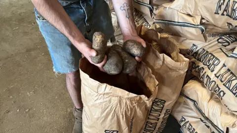 John Devine/BBC Mr Ablett's arms are visible as he shows off three large potatoes from an open brown paper sack, he has Demin shorts and brown ankle boots.