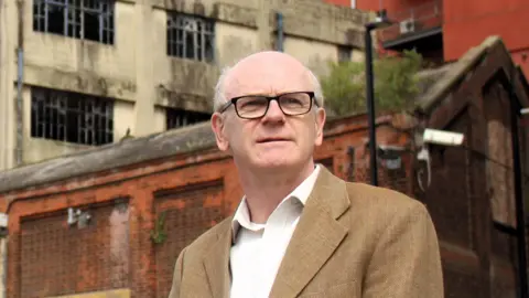 Ipswich Borough Council Neil MacDonald, Ipswich's Labour leader is wearing a light brown blazer and a white shirt. He is wearing black frame glasses and appears to be looking beyond the camera.
The camera appears to be positioned slightly below MacDonald and looking up at him. 