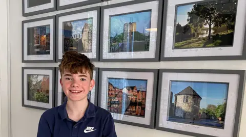 Isaac stood in front of a white wall with eight picture frames behind him with his photography in them. All the pictures have black frames in two rows of four. Isaac has short brown hair and is wearing a navy blue polo t-shirt.