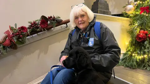 A woman sat at the top of a staircase next to a green decorated Christmas tree. She has short white hair with blue glasses resting on top of her head. She is sitting next to a small, black labrador puppy and wearing a black jacket with a badge that reads 'Guide Dogs'.