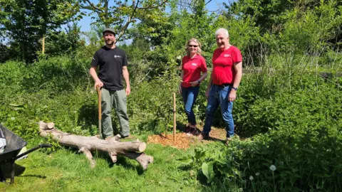 A woman and a man wearing red t-shirts and blue jeans stand near an area of vegetation while a man in a black top with green trousers stands next to a log while holding a spade. 