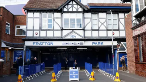 A generic photo taken previously shows blue barriers lined up outside the entrance to Fratton Park, with Fratton Park written in blue above on the front of the building