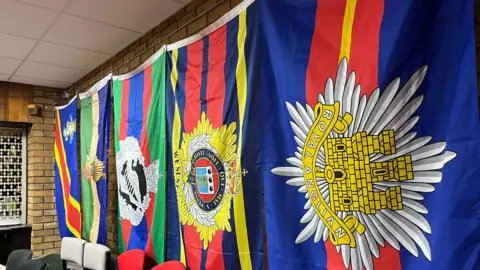 Tom Jackson/ BBC A row of brightly colour flags have been pinned against a brick wall inside the club . The closest to the camera reads 'Royal Anglian'. 