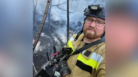 Ed Matthews, who wears a protective uniform, holds a camera. He is stood in a smoky woodland area.