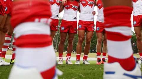 A pair of blurry legs in the foreground with a line of Sydney Swans players in the background