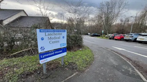 A sign reading Lanchester Medical Centre on the side of a pavement leading to a car park.
