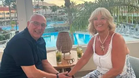 supplied A middle age couple are sitting side on to the camera. He is holding her right hand. They are at a holiday destination, with a pool in the background and are smiling at the camera. He is wearing a navy shirt and she has a white sleveless top 