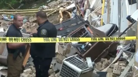 Two police officers stand behind yellow police tape looking at a destroyed apartment complex.